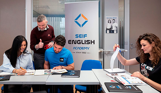 Fotografía de una clase presencial de nuestros cursos de inglés en Madrid. Un profesor, Blake, observa lo escrito por dos alumnos en sus libros, e indica que la respuesta es correcta con el pulgar hacia arriba. La escena tiene lugar en un aula de nuestra academia de inglés en Gran Vía 50.
