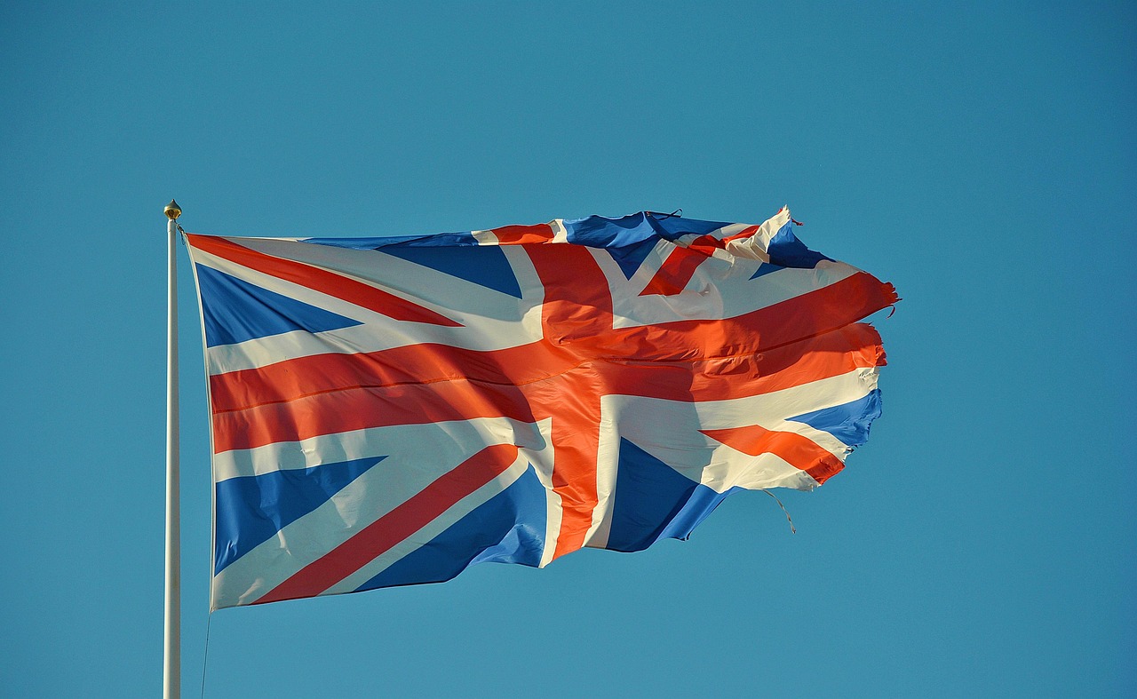 Bandera británica ondeando en un cielo azul, con el texto «Examen Aptis, diferencias entre Curso A2-B2 y Curso B2-C1»